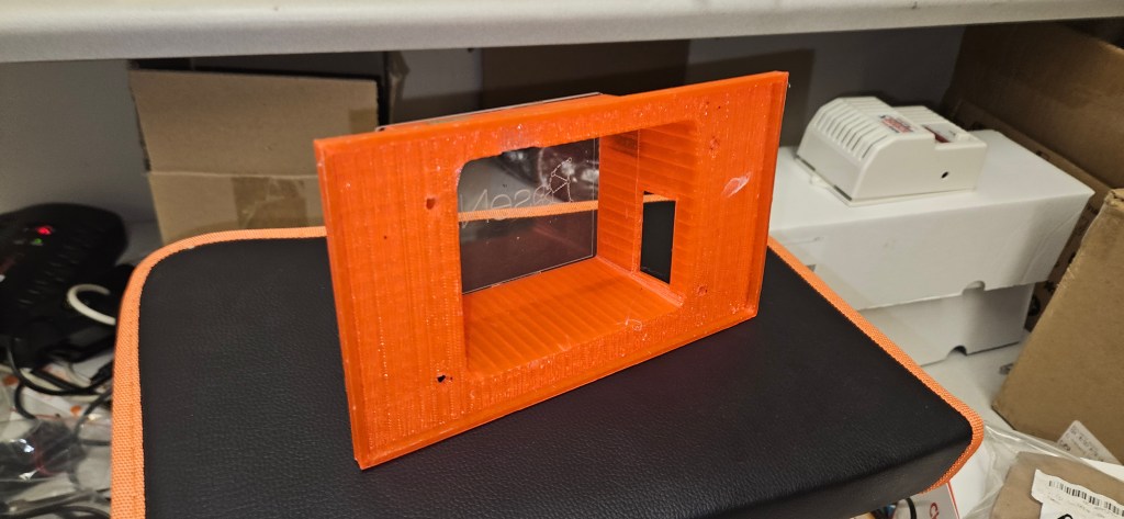 The image shows a small orange plastic object with a window on top of a black surface, possibly a table or desk. The objec...