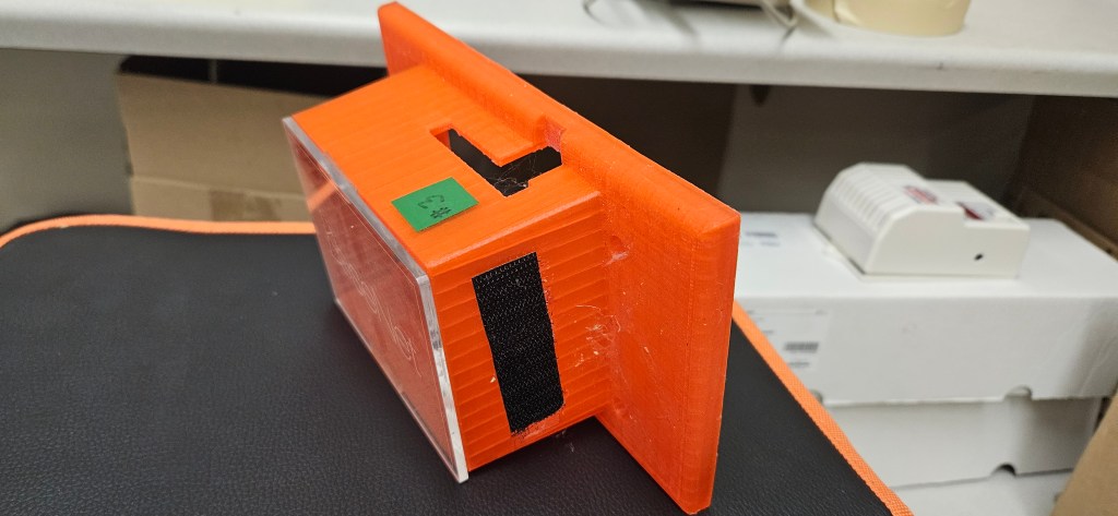 An orange 3D printed object, possibly a prototype, sits on a black mat with an orange border. The object features a green ...