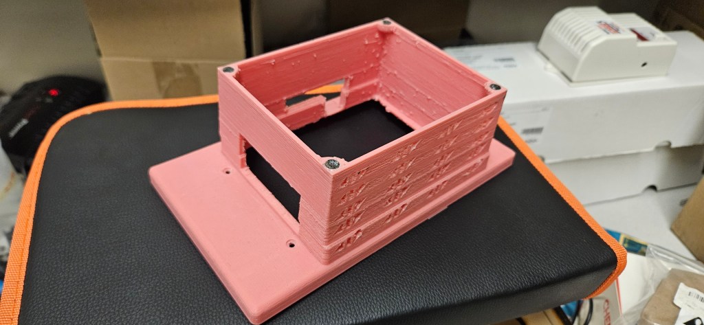 A 3D printed object sits atop a black and orange cushion, with a white box and cardboard box in the background.