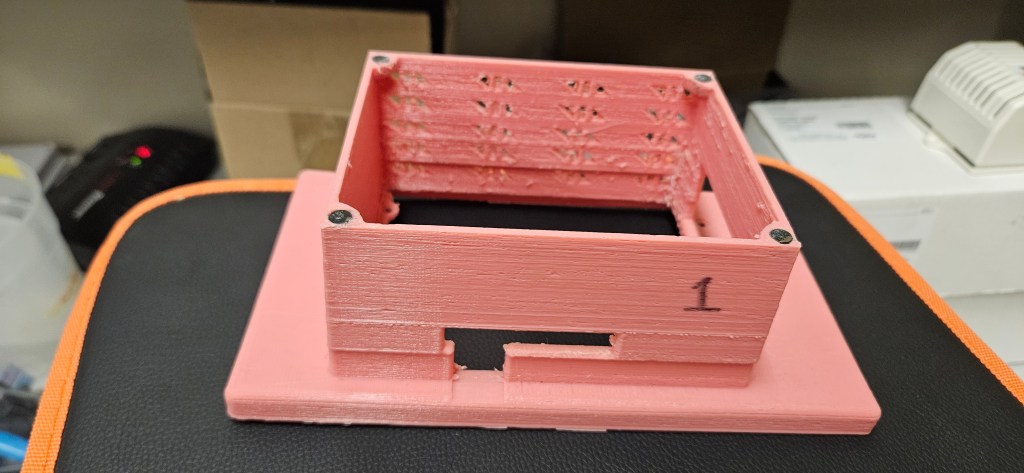 A pink 3D printed object sits on a black surface with orange trim, possibly a table or chair. The object appears to be a b...