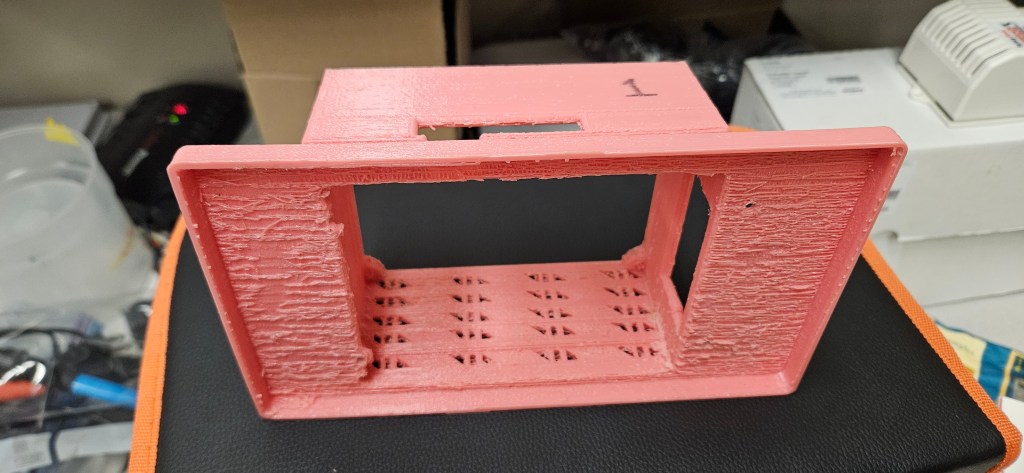 A pink 3D printed object with a rectangular shape and square holes sits atop a black and orange surface, with a white box ...