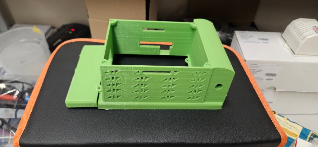 The image shows a 3D printed object in green on top of a black and orange case. The object appears to be a small box or co...