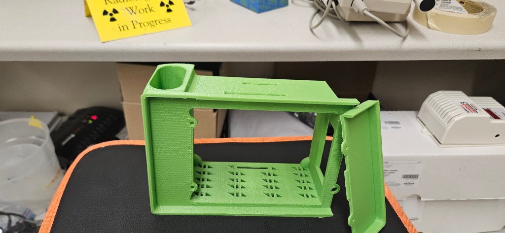 A green 3D printed object sits on a black and orange chair, with a white desk and various objects in the background. The o...