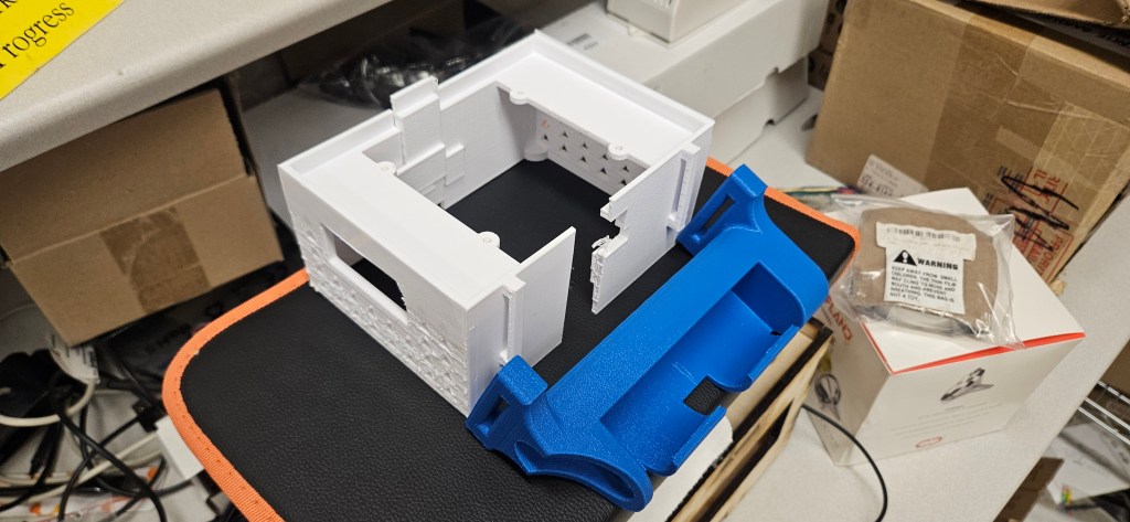 A 3D printed object, likely a house or building, sits on a black and orange mat in a cluttered room. The object is white w...