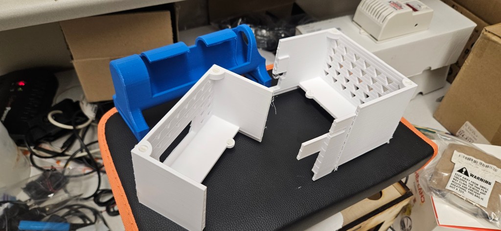 A 3D printed object sits on a black and orange laptop case, surrounded by a cluttered workspace with various cords, boxes,...