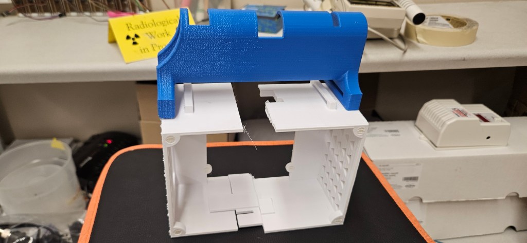 A 3D printed object sits atop a black and orange table, surrounded by various scientific equipment and supplies in the bac...