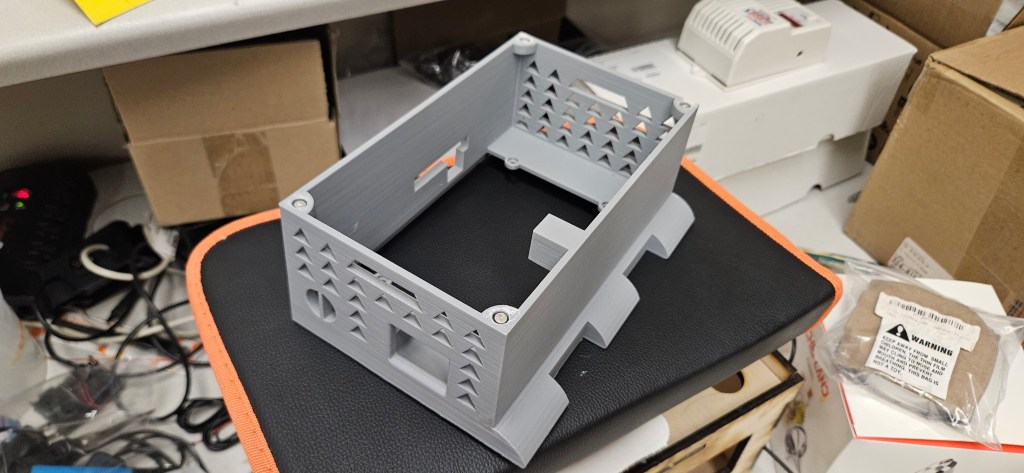 A 3D printed gray plastic box with a rectangular shape and a square base, sitting on a black and orange surface.