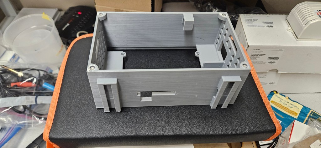 A gray plastic 3D printed box on top of a black and orange case, surrounded by various items in the background.