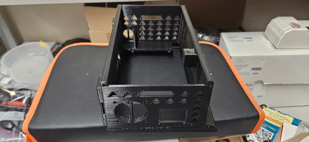 A 3D printed object sits atop an orange and black case, surrounded by various tools and supplies in a cluttered workshop s...