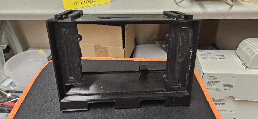 A black plastic object sits on a gray and orange mat, surrounded by boxes and tools. The object appears to be a 3D printed...