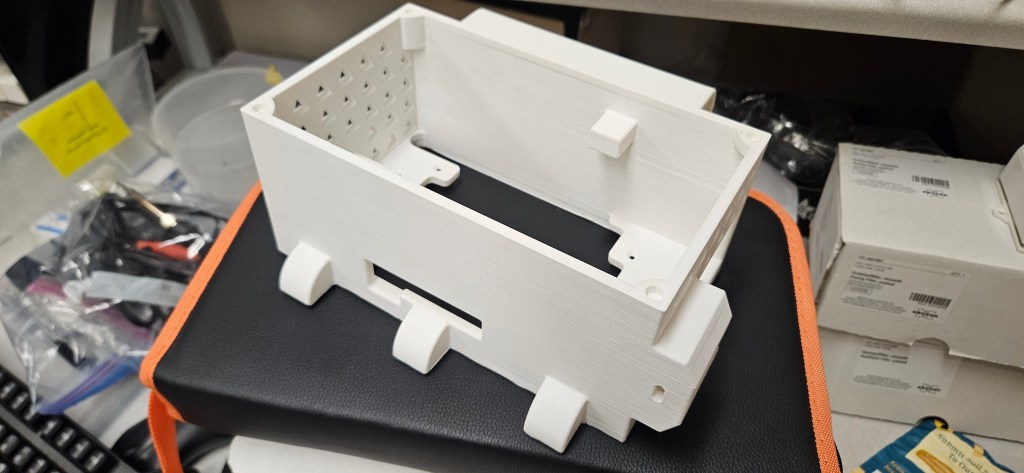 A white 3D printed object sits on a black and orange surface, surrounded by various items.