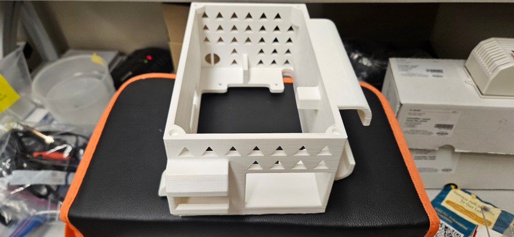 A white 3D printed object sits on top of a black and orange case in an office setting.