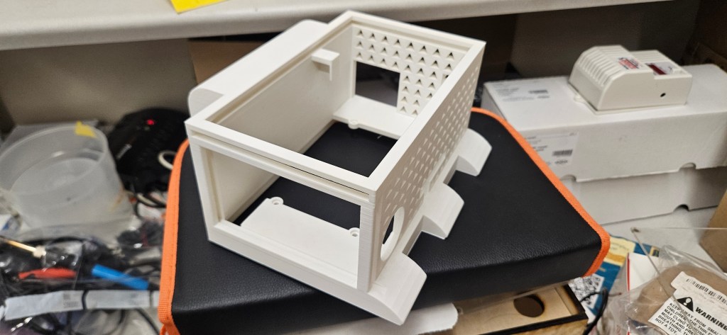 A 3D printed object sits atop a black and orange case, surrounded by various electronic components and tools on a cluttere...