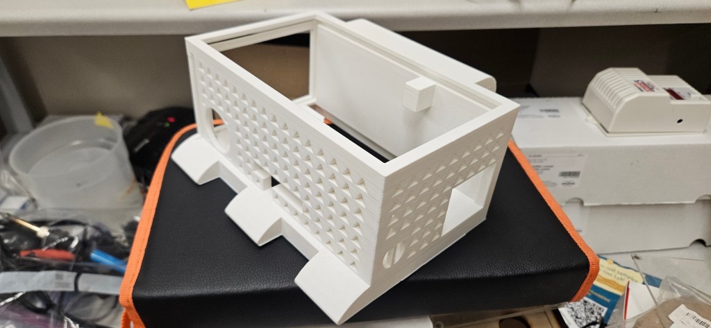 A white 3D-printed object with a rectangular shape and square holes sits atop a black box with orange trim, surrounded by ...