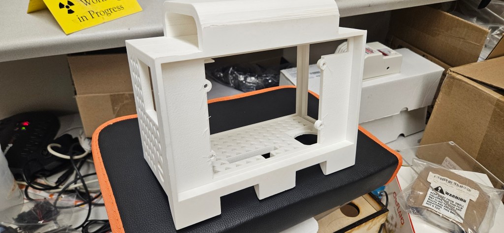 A white 3D printed object sits atop a black and orange mat, surrounded by cardboard boxes, cords, and other electronics. T...