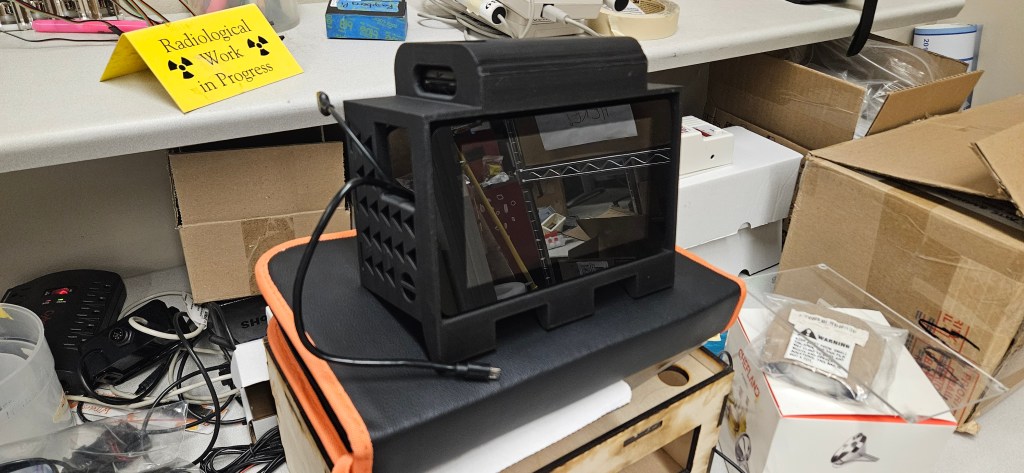 A 3D printer sits atop a black and orange case, surrounded by various items on a cluttered table. The printer itself is bl...