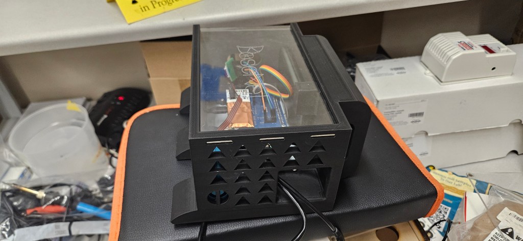 A cluttered workspace with a black device featuring a clear plastic top and a collection of wires and cables inside, place...