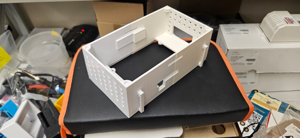 A white 3D printed object with a rectangular shape and holes sits atop a black case with an orange strap, surrounded by va...