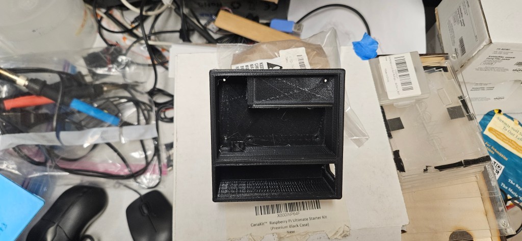 A 3D-printed object sits on a white surface amidst a cluttered background of various electronic components and tools.