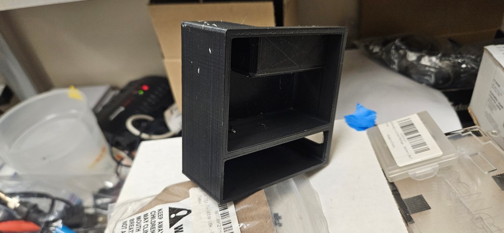 A black 3D-printed object sits on a white surface, surrounded by various items in the background. The object has a rectang...