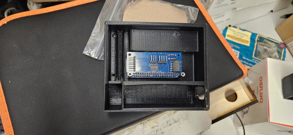 A black plastic box with an open top, containing a blue circuit board and a small chip, placed on a black and orange surface.