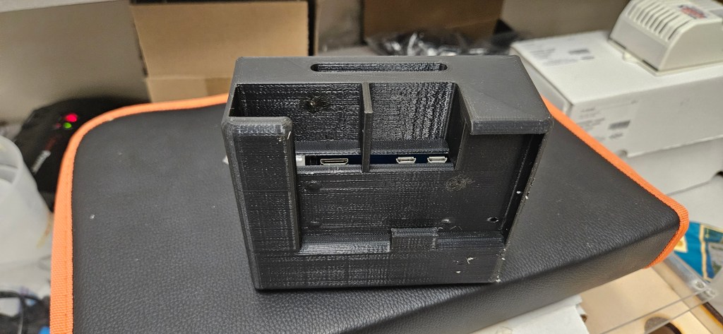 A 3D-printed black plastic device with a rectangular shape, featuring two USB ports and a small hole on top, placed on a b...