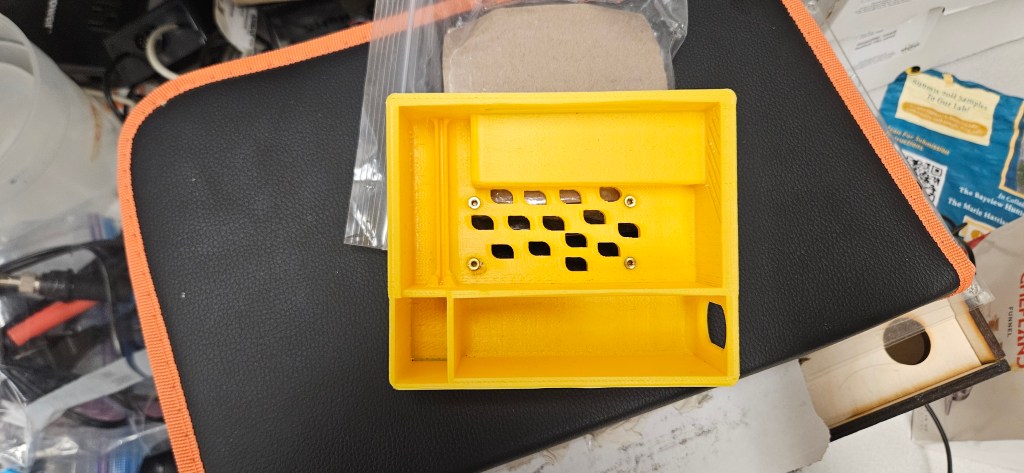 A yellow 3D-printed object with holes in it, placed on top of a black and orange case, possibly for a Raspberry Pi or othe...