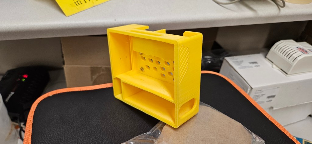 A yellow 3D printed object sits atop a black chair with orange trim, positioned in front of a white desk and surrounded by...